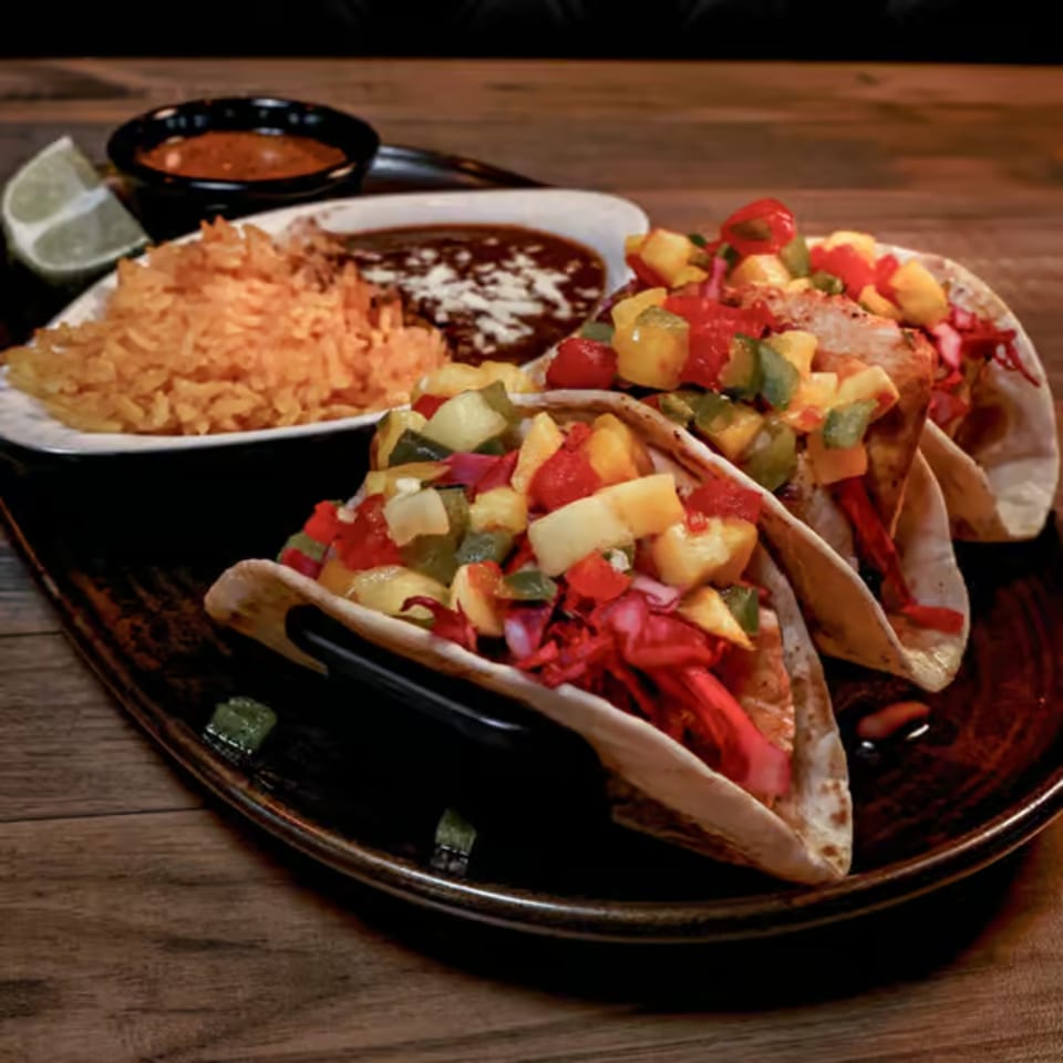 Three vibrant Santo Tequila tacos with colorful toppings, rice, beans, and a lime wedge on a wooden table.