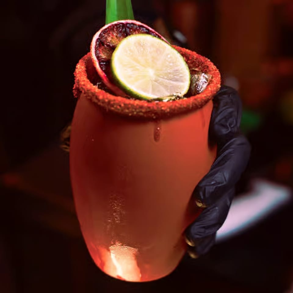 A hand in a black glove holds a red cup with a lime and orange slice, rimmed with chili powder.