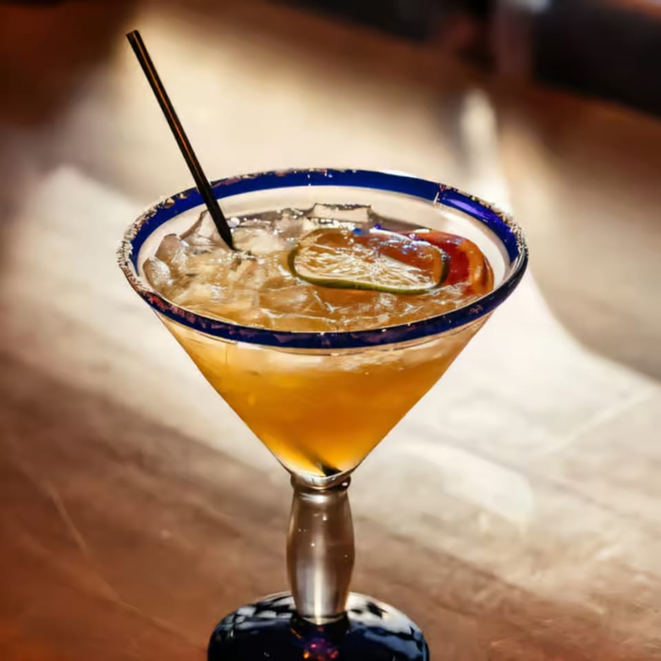 A Santo Tequila margarita in a blue-rimmed glass with ice, lime, and a straw.
