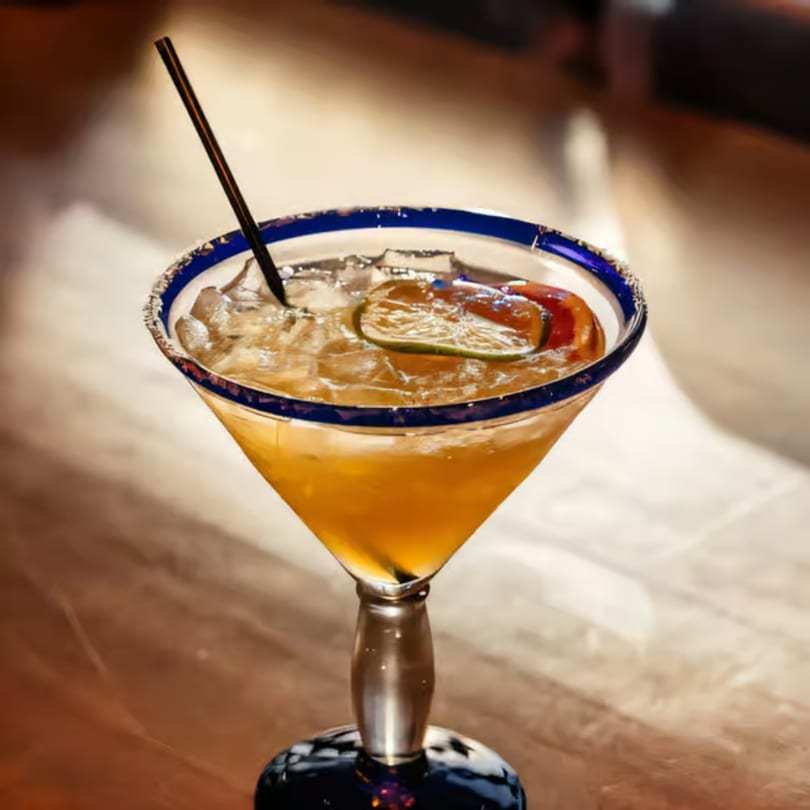 A Santo Tequila margarita in a blue-rimmed glass with ice, lime, and a straw.
