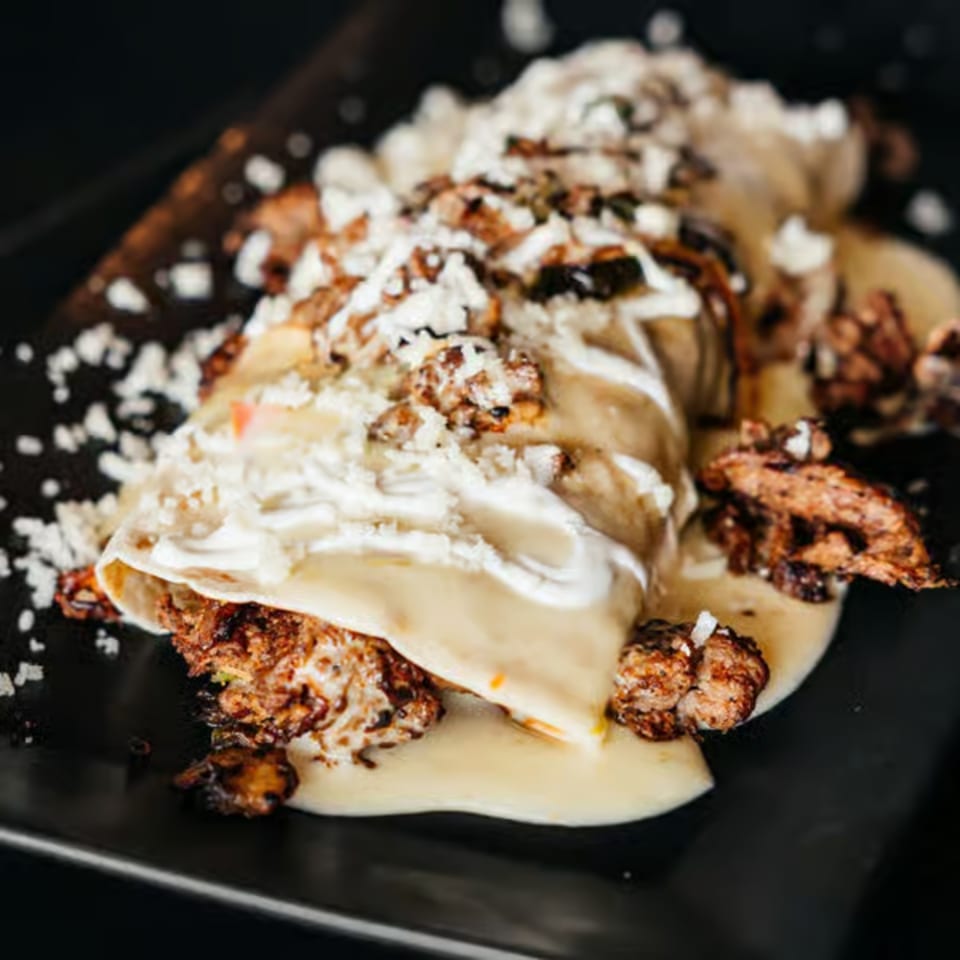 A close-up of a Santo Tequila Mexican enchilada, topped with white sauce, crumbled cheese, and meat.